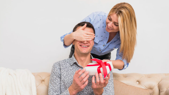 Женщина закрыла рукой глаза сидячему мужчине на диване, дарит подарок, мужчина и женщина улыбаются