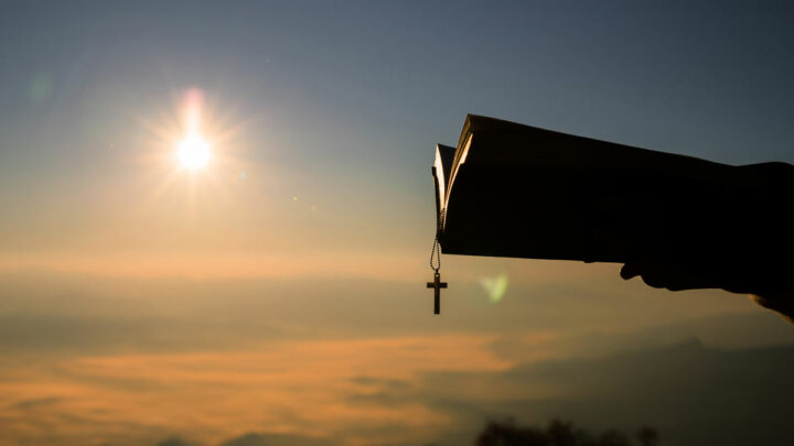 силуэт человека, держащего библию и крест, фон - восход солнца