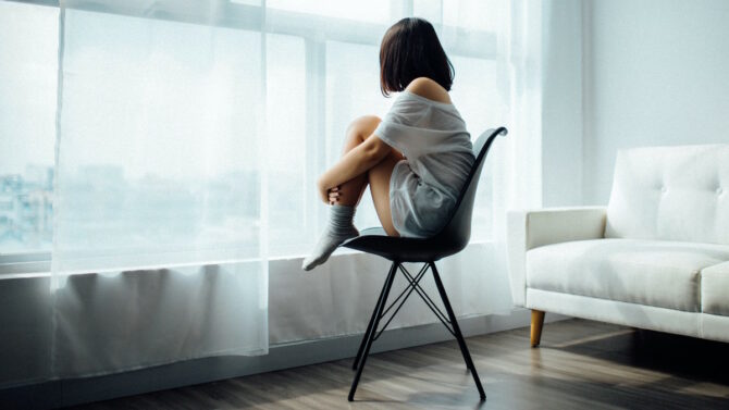 Девушка сидит у окна на стуле обхватив колени