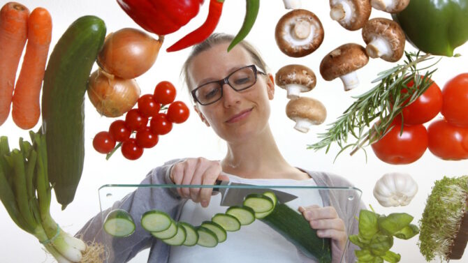 Женщина в окружении овощей режет огурец